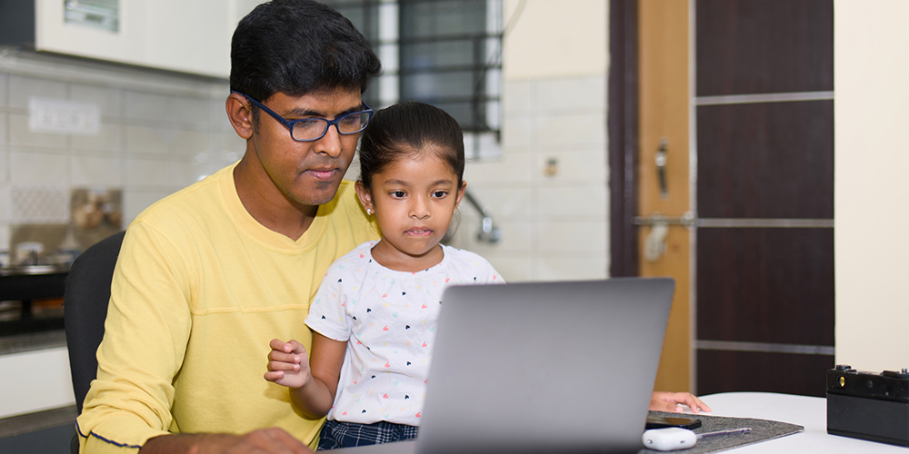 A man wearing glasses and a yellow shirt sits at a table with a young girl on his lap, both focused on a laptop screen in a home setting